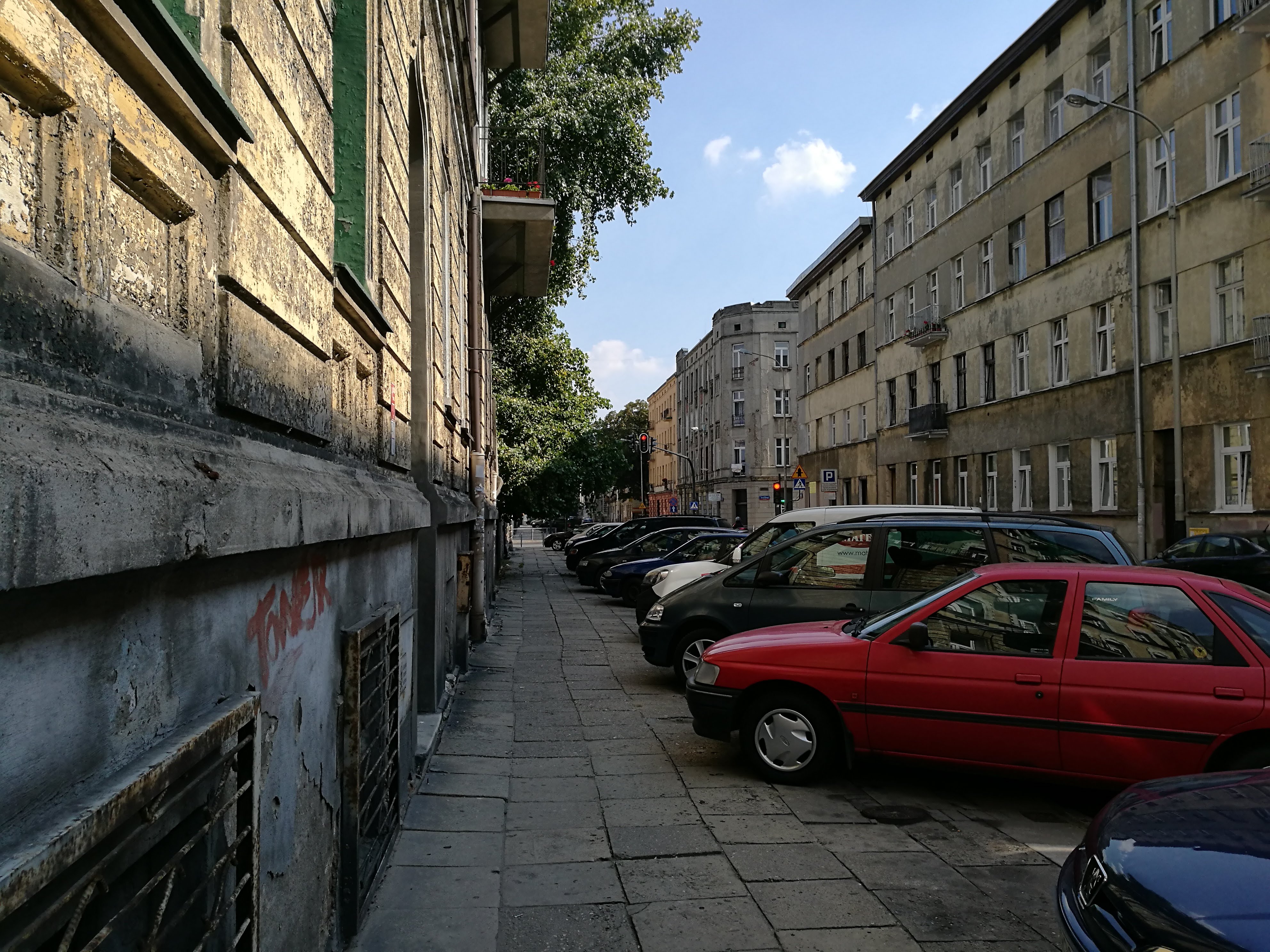 A row of cars parked up on the pavement, covering most of the path. It's a city centre street surrounded by tall apartment buildings.